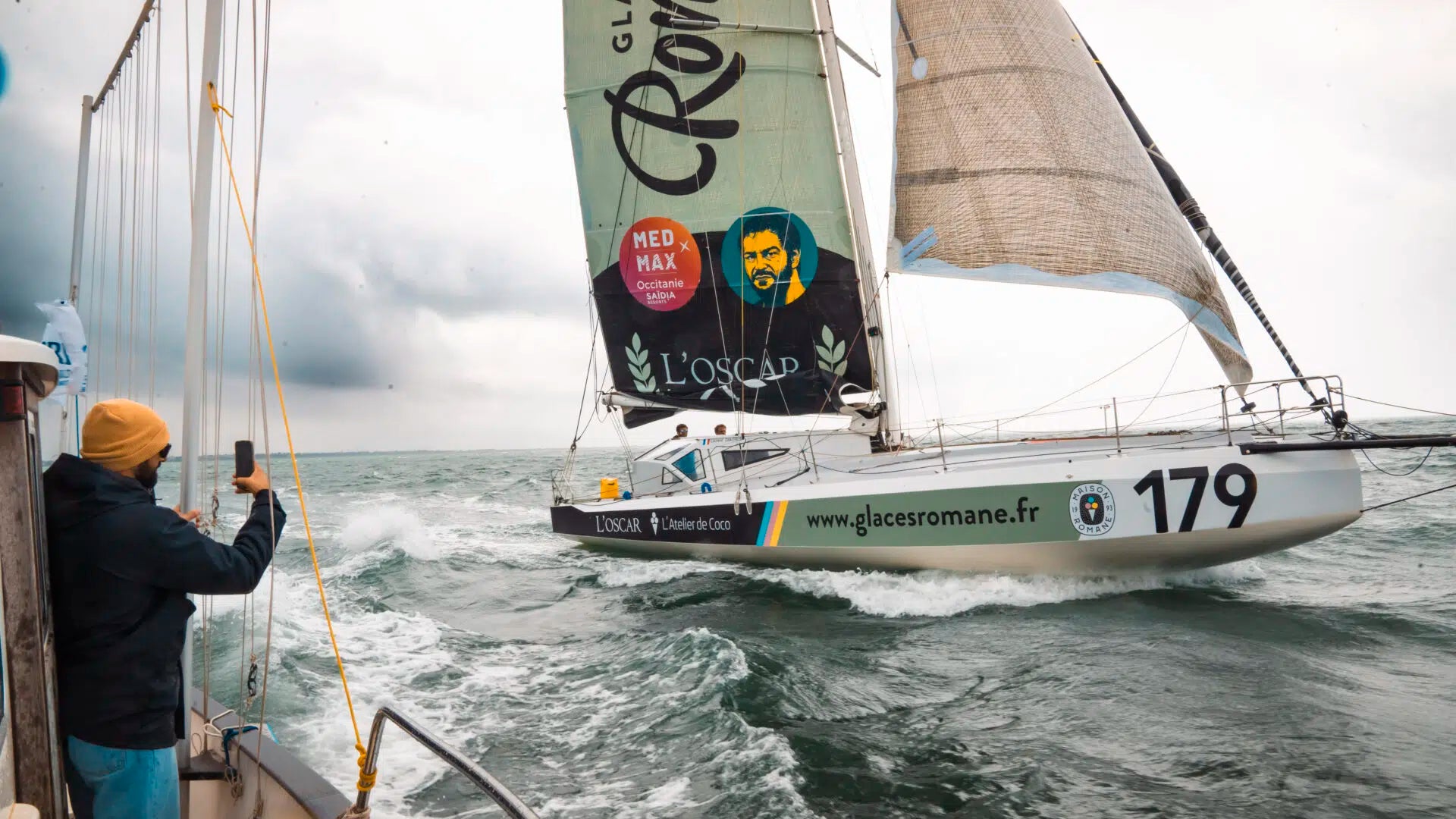 Class 40 #179 Glaces Romane (Laurent Camprubi) en navigation, voile portant le portrait du skipper et les sponsors L'Oscar / Med Max Occitanie